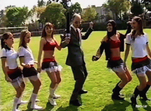 Cheerleaders Dancing On Field With Suited Man Holding Gun GIF