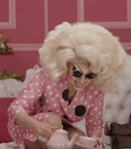 Drag Queen With Blonde Wig And Teapot On Bed GIF