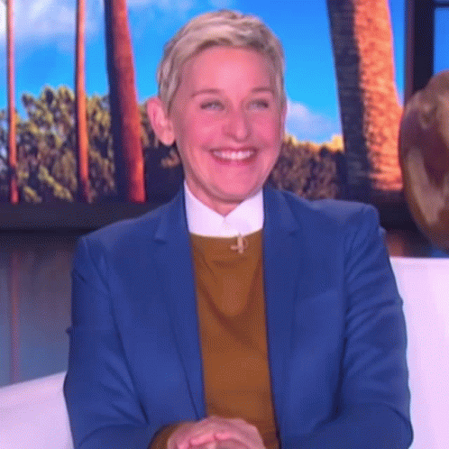 Ellen Degeneres Is Wearing A Blue Suit And A Yellow Sweater And Smiling While Sitting On A Couch . GIF