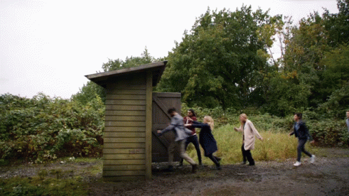 Group Standing Around Wooden Outhouse GIF