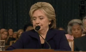 Hillary Clinton Is Speaking Into A Microphone While Sitting At A Desk In Front Of A Crowd . GIF