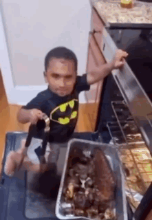 Little Boy in Batman Shirt Standing by Oven With Pan of Food