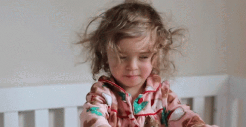 Little Girl With Curly Hair Sitting in Crib Looking at Camera