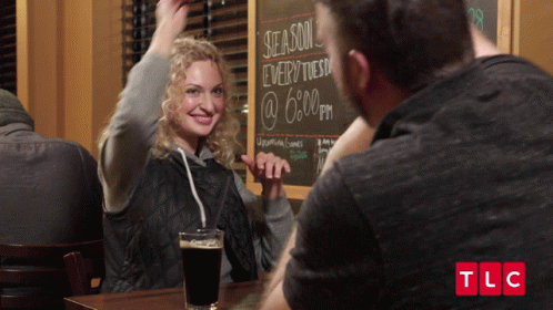 Man And Woman Sitting At Table With Chalkboard Saying Season Every Tuesday @ 6 PM GIF