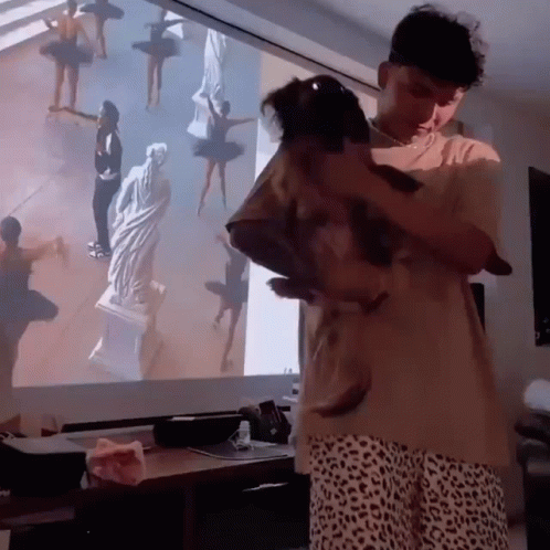 Man Holding Small Dog in Front of Projector Screen With Ballerinas