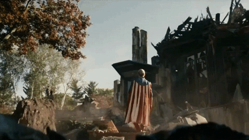 Man in American Flag Cape Standing by Destroyed House
