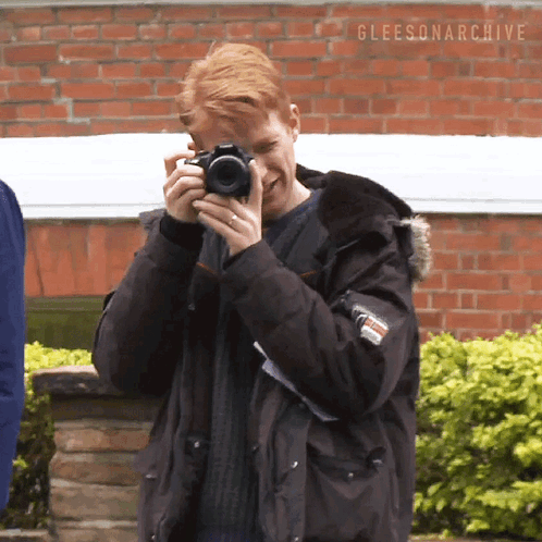 Man In Black Jacket Holding Camera In Front Of Brick Wall Gleeson Archive GIF