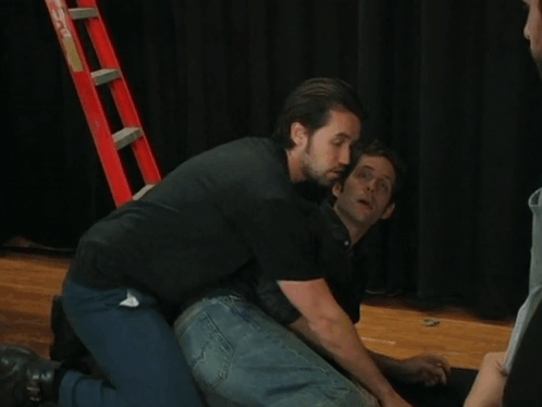 Man In Black Shirt Kneeling Next To Man In Blue Jeans GIF