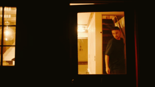 Man in Black Shirt Looking Out Window in Dark Room
