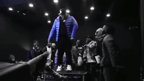 Man In Blue Jacket Standing On Table In Front Of Crowd GIF