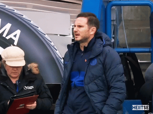 Man In Blue Jacket Stands In Front Of Heitse Sign GIF