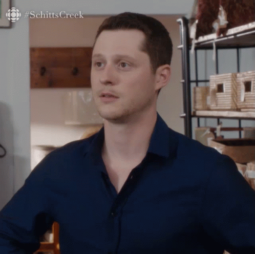 Man In Blue Shirt In Front Of Schitts Creek Shelf GIF