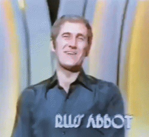 Man In Blue Shirt Smiling In Front Of Rus Abbot Sign GIF