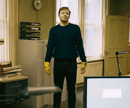 Man In Blue Sweater And Yellow Gloves Standing Before Clock GIF