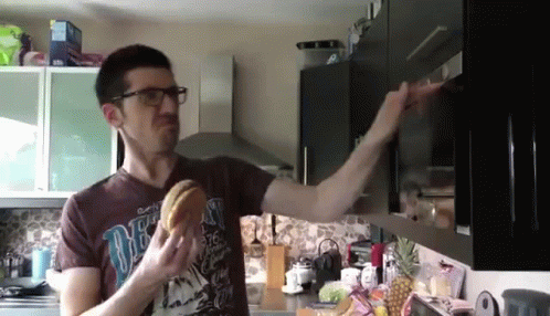 Man in Brown Shirt Saying One Eating Hamburger in Kitchen
