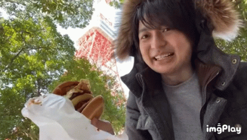 Man In Fur Hood Holding Hamburger In Front Of Tower And Trees GIF