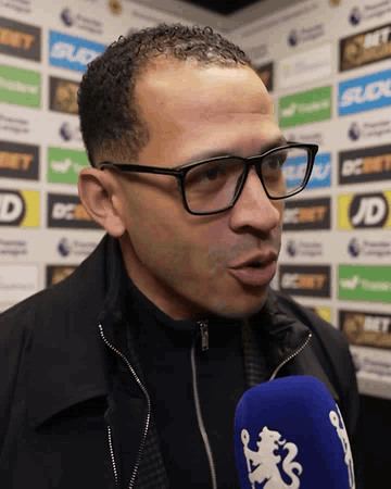 Man in Glasses and Black Jacket Talking Into Blue Microphone