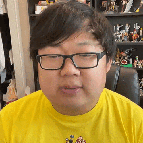 Man in Glasses and Yellow Shirt Sitting in Front of Toy Shelf