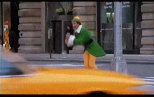 Man In Green Elf Costume Walking Next To Yellow Taxi GIF