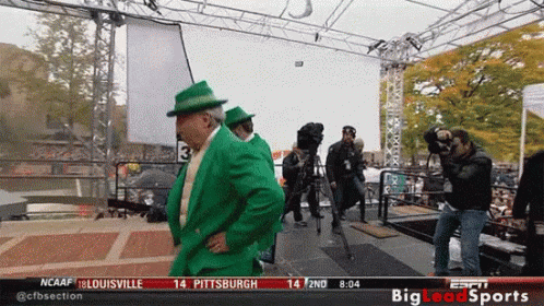 Man in Green Suit and Hat in Front of NCAAF Sign