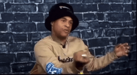 Man In Hat And Sweater Sitting By Brick Wall Talking GIF