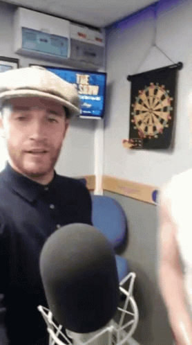 Man In Hat At Microphone In Front Of Dart Board GIF