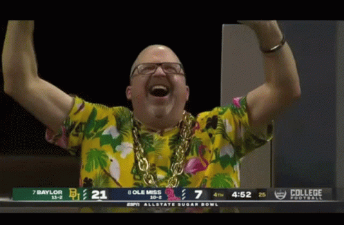 Man in Hawaiian Shirt and Gold Chain in Front of College Football Scoreboard