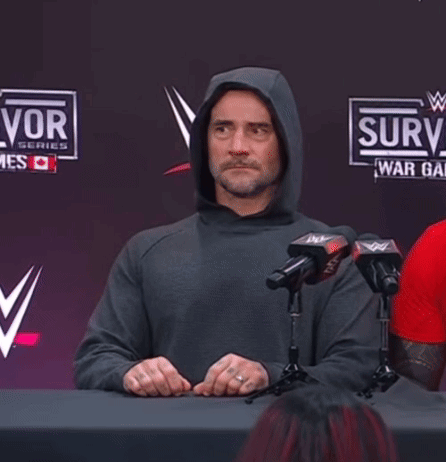 Man in Hoodie Sitting at Table With Microphones in Front of Surv Sign