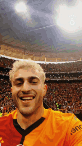 Man In Orange Shirt Saying Eme Smiling In Crowd GIF