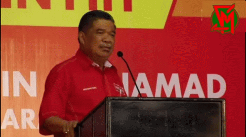Man in Red Shirt Speaking at Podium In Jamad