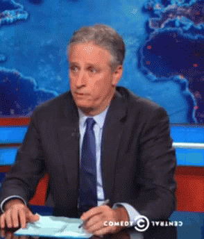 Man In Suit Sitting At Desk In Front Of Comedy Central Screen GIF