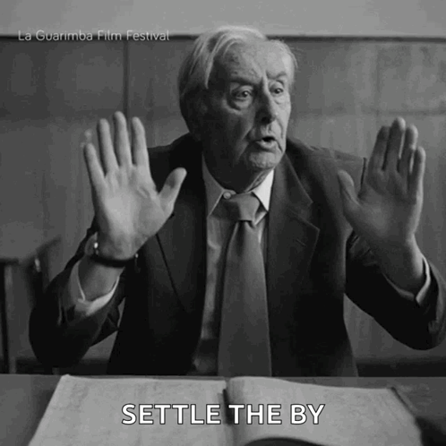 Man In Suit Sitting At Desk With Hands Up Saying Settle GIF