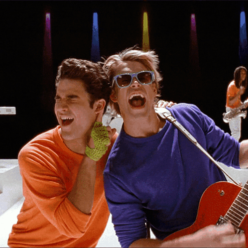 Man in Sunglasses Singing Into Microphone Beside Man in Orange Shirt