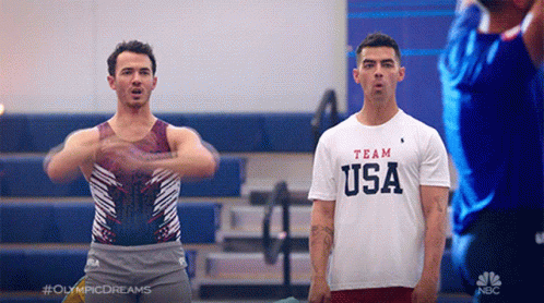 Man In Team USA Shirt Standing Next To Man In Leotard GIF