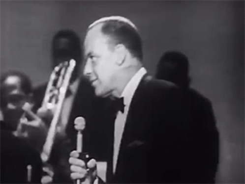 Man in Tuxedo Singing Into Microphone With Band Behind