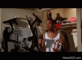 Man in White Tank Top Standing Surrounded by Guns