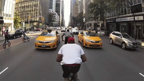 Man Riding Bike Down Street With Taxis and Bank of America