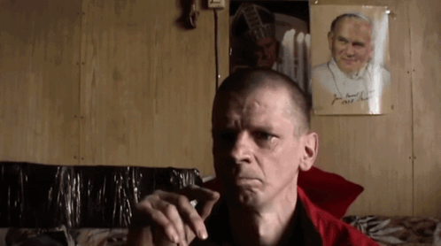 Man Smoking Cigarette In Front Of Picture Named John GIF