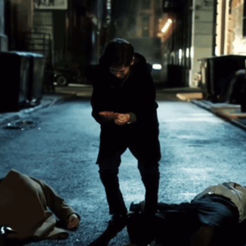 Man Standing Next to Man Laying on Ground in Dark Alleyway