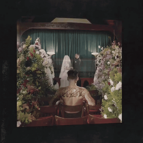 Man With Back Tattoo Beside Coffin and Flowers