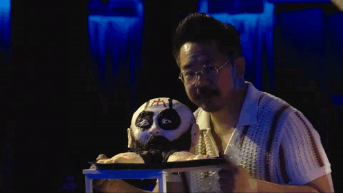 Man With Glasses Holding Puppet With Cross on Face