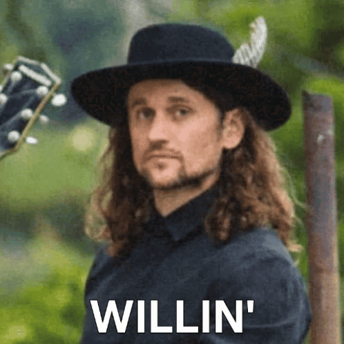 Man With Long Hair in Black Hat Holding Guitar 'Willin