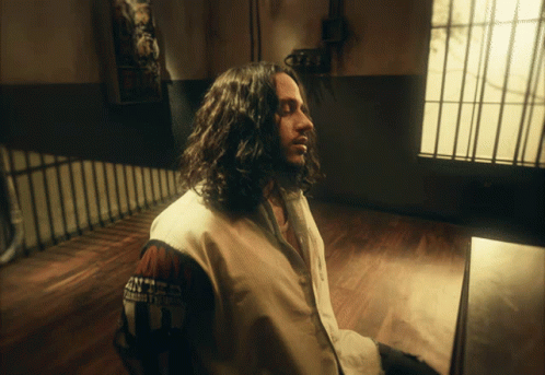 Man With Long Hair Sitting In Room Wearing U.S. Army Jacket GIF