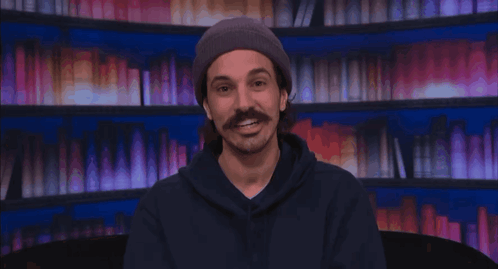 Man With Mustache in Beanie and Hoodie Smiling by Bookshelf