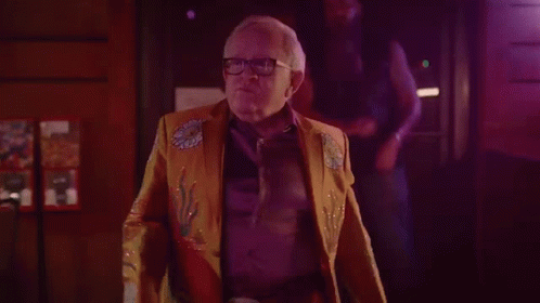 Older Man In Yellow Jacket And Glasses In Dark Room GIF