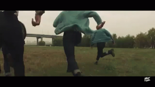 People Running Through Grassy Field in Front of Bridge