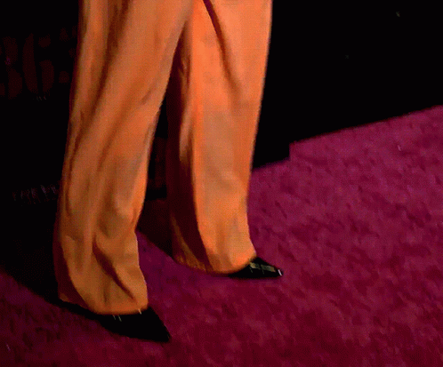 Person in Orange Pants and Black Shoes Walking on Pink Carpet