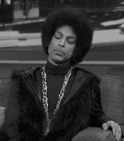 Prince Is Sitting On A Couch Wearing A Fur Vest And A Chain Around His Neck . GIF