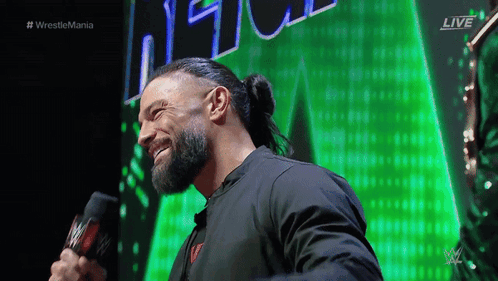 Roman Reigns Is Smiling While Holding A Microphone In Front Of A Green Background GIF