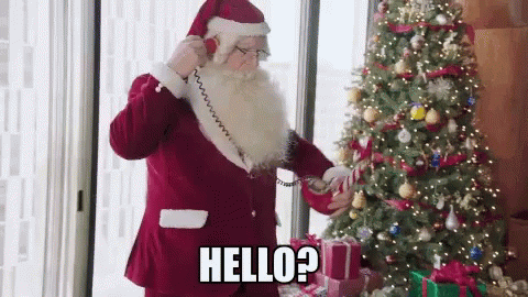 Santa Claus Is Talking On A Phone In Front Of A Christmas Tree And Says Hello ? GIF